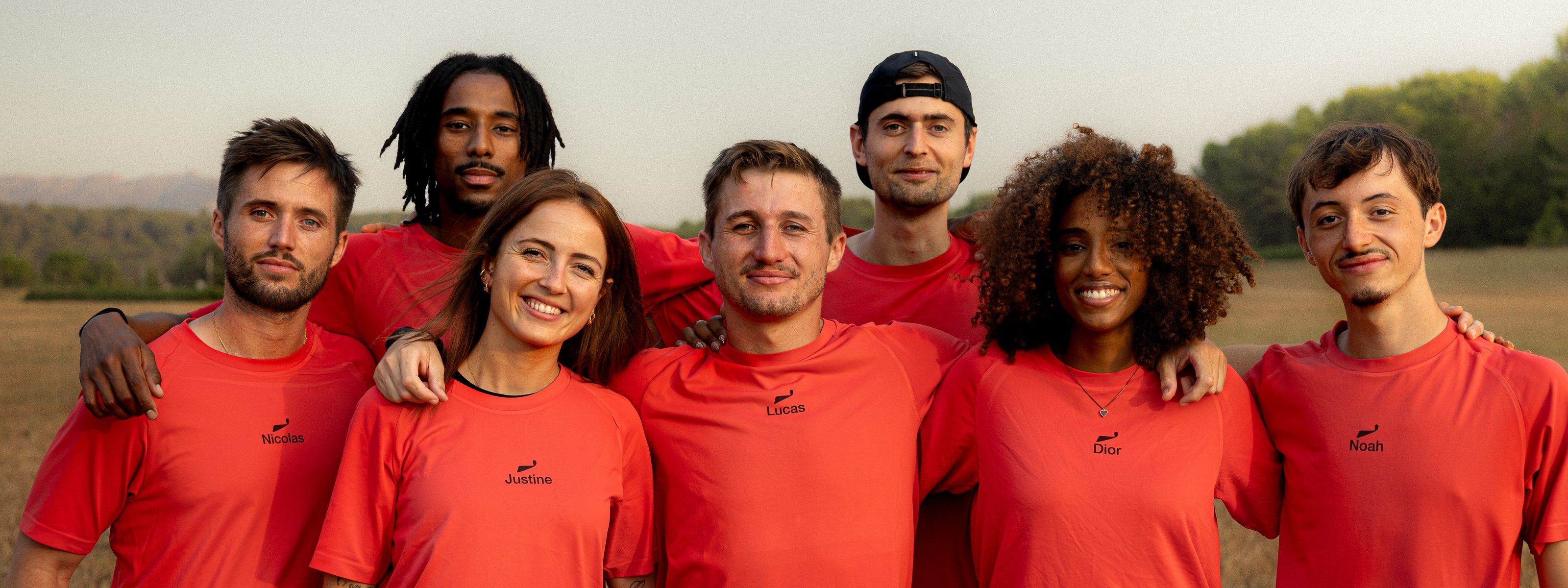 Sept membres du staff, bras dessus bras dessous dans un champ, en T-shirts rouges WAVE ; sous la vague brodée, on lit les prénoms Nicolas, Justine, Lucas, Dior et Noah.